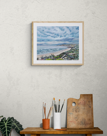 Framed artwork of a moody, South Australian, coastal scene on a wall above a wooden table with art supplies.